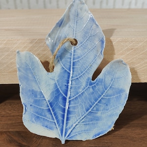 Może przedstawiać: Niebieski ceramiczny ornament w kształcie liścia z białym szkliwem i szczegółowym wzorem żyłek. Liść ma otwór na górze do zawieszenia.