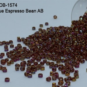 May include: A collection of Opaque Espresso Bean AB beads, with a mix of brown, gold, and purple hues, spilling from a clear container. The beads are cylindrical and scattered across a light surface. Text on the image reads "Item: DB-1574 Opaque Espresso Bean AB".