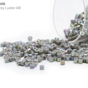 Könnte beinhalten: Nahaufnahme eines Haufens kleiner, runder, grauer Perlen mit irisierendem Finish. Die Perlen fallen aus einem durchsichtigen Glasbehälter. Die Perlen sind mit "DB-0168 Opaque Grey Luster AB" gekennzeichnet.