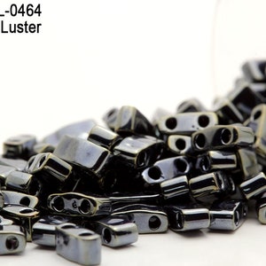 Pode incluir: Um close-up de um monte de contas de vidro cinza escuro com brilho. As contas são retangulares com dois furos e têm um brilho metálico.