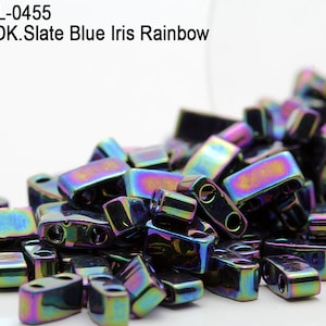 Pode incluir: Um close-up de um monte de contas de vidro iridescentes de cor arco-íris azul ardósia escuro. As contas são retangulares com dois furos e estão a cair de um frasco de vidro transparente.