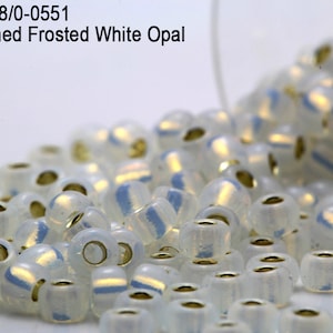 May include: A close-up of a pile of small, white, frosted glass beads with silver lining. The beads have a slight iridescent sheen. Item: RR8/0-0551 Silver Lined Frosted White Opal