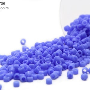 May include: A close-up of a container of opaque sapphire blue seed beads. The beads are small and round, and they are spilling out of the container onto a white surface. Item: DB-0730