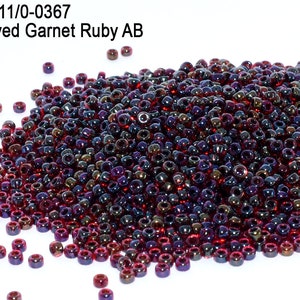 May include: A pile of small, round, red and purple glass beads with an iridescent finish. The beads are labeled "Inside Dyed Garnet Ruby AB".