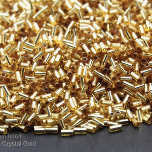 May include: A close-up image of a pile of gold-colored glass beads. The beads are small and cylindrical, with a silver lining. The beads are arranged in a random pattern, creating a shimmering effect. Item: RB 3mm-0004 Silver Lined Crystal Gold