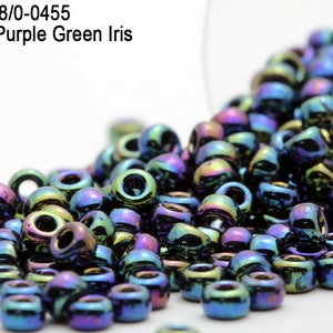 Op de afbeelding: Een verzameling metallic paarse en groene iris kralen, verspreid uit een doorzichtige container. De kralen zijn cilindrisch met een gat in het midden. De tekst luidt "Item: RR8/0-0455 Metallic Purple Green Iris".