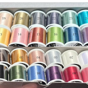 May include: A selection of colourful thread spools in a box. The threads are in various shades, including pink, gold, blue, green, and purple. Each spool is labelled with a number.