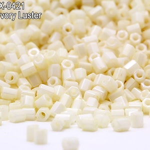 May include: Close-up of a pile of opaque ivory luster beads. The beads are hexagonal in shape and have a slightly glossy finish. The text "Item: HEX-0421 Opaque Ivory Luster" is visible in the upper left corner. These beads are ideal for jewelry making.