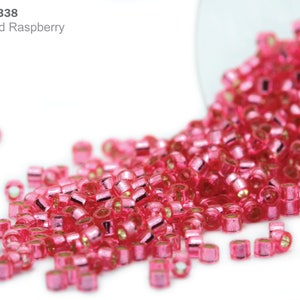 May include: A close-up of a pile of pink seed beads with a silver lining. The beads are spilling out of a clear glass container.