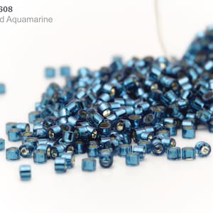 May include: A close-up of a pile of small, blue, metallic beads. The beads are round and have a shiny finish. The beads are scattered on a white surface. Item: DB-0608 Inside Dyed Aquamarine