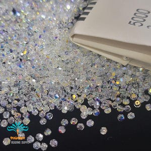 Puede incluir: Primer plano de una gran cantidad de cuentas de vidrio transparentes. Las cuentas son pequeñas y tienen un acabado facetado e iridiscente. Las cuentas están dispuestas en un montón suelto sobre una superficie negra. Una etiqueta blanca con el texto "5000 2 mm" es visible en el fondo.