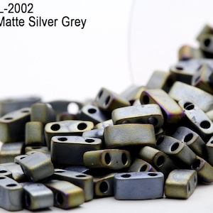 Puede incluir: Primer plano de un montón de cuentas metálicas mate de color gris plateado. Las cuentas son de forma rectangular y tienen un acabado ligeramente iridiscente.