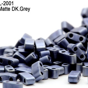 Puede incluir: Primer plano de un montón de cuentas de dos agujeros de color gris oscuro mate metálico. Las cuentas están en un recipiente transparente y se están derramando sobre una superficie blanca. El texto "Item: HTL-2001 Metallic Matte DK.Grey" es visible en la parte superior de la imagen.