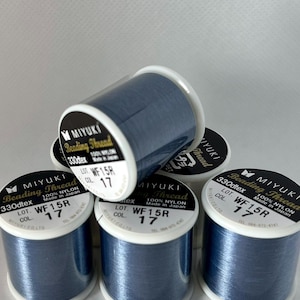May include: Several spools of Miyuki Beading Thread in a deep blue color. Each spool has a white label with black text indicating the thread type, lot number, and that it is made of 100% nylon. The spools are stacked and arranged.