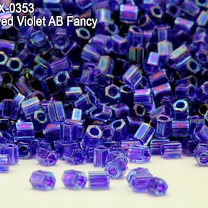 May include: A close-up image of a pile of small, iridescent violet hexagonal beads. The beads have a shiny, reflective finish and are arranged in a random pattern. The beads are labeled "HEX-0353 Inside Dyed Violet AB Fancy".