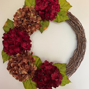 May include: A grapevine wreath adorned with burgundy and brown hydrangea blooms and green leaves. The wreath is a circular shape, perfect for seasonal home decor. The flowers are clustered together, creating a full and vibrant look.