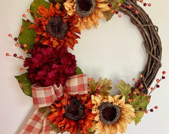 May include: A fall-themed wreath with a brown vine base, decorated with sunflowers, burgundy hydrangeas, and red berries. A red and white checkered ribbon is tied around the wreath.