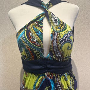May include: A sleeveless dress featuring a vibrant paisley print in shades of lime green, navy blue, orange, and turquoise. The bodice has a twisted halter neckline with a deep cutout and a solid navy blue ruched waistband that sits above the skirt.