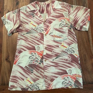 May include: A short-sleeve button-down shirt featuring a maroon and cream color palette with a repeating pattern of Japanese-style folding fans, pagodas, bridges, and circular motifs. The shirt has a classic collar and a single chest pocket.