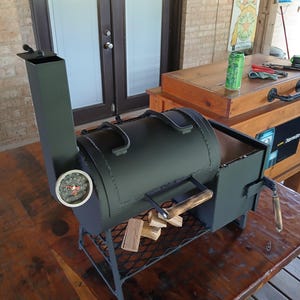 May include: A black metal smoker with a round body, a chimney, and a temperature gauge. It has a handle and sits on a metal stand. Wood pieces are placed on a lower rack. The smoker is on a wooden table.