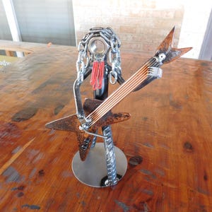 May include: A metal sculpture of a musician playing a guitar. The sculpture is made of various metal pieces, including chains, rods, and a circular base. The guitar has copper-colored strings and a star-shaped body. The musician has a chain-like head and red accents.