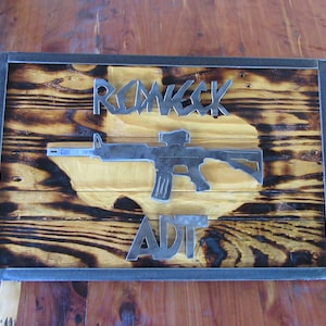 May include: A wooden sign with a metal frame and a burnt wood finish. The sign features a metal cutout of a rifle with a scope, the words "Redneck" and "ADT".