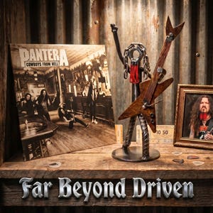 May include: A display shelf featuring a Pantera album cover, a metal sculpture of a guitarist, and a framed photo. The album is titled "Cowboys From Hell". The sculpture has a guitar-shaped body. The text "Far Beyond Driven" is at the bottom.
