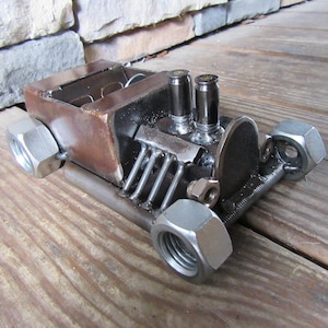May include: A small, handmade model car made from metal nuts, bolts, and other scrap metal. The car is a hot rod style with a large engine and two exhaust pipes. The car is painted in a rusty brown color.