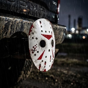 May include: A white hockey mask with red accents and blood-like splatters is mounted on the back of a truck. The mask has several holes and is set against a dark, rainy backdrop. The mask is a horror movie prop.