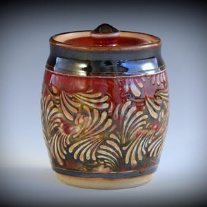 May include: A handcrafted ceramic jar with a lid. The jar is cylindrical with a reddish-brown glaze and intricate, swirling floral patterns in cream and gold. The rim and lid are dark brown.