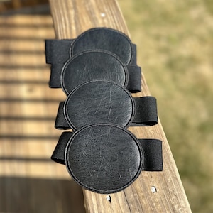 Może przedstawiać: Cztery czarne, okrągłe podkładki pod szklanki z teksturowaną powierzchnią i elastycznymi paskami. Podkładki ułożone są w rzędzie na drewnianej powierzchni, prezentując prosty i funkcjonalny design. Chronią powierzchnie przed śladami po wodzie.