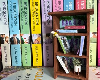 May include: A brown wooden bookshelf with three shelves. The shelves are filled with books, including a few with the title "Bridgerton" on the spine. There is a small potted plant on the top shelf.