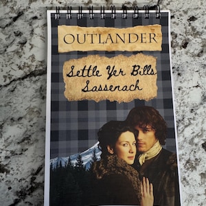 May include: Spiral-bound notepad featuring the word "OUTLANDER" and the phrase "Settle Yer Bills Sassenach" on a plaid background. The notepad includes an image of two people in period clothing, with a mountain and forest backdrop.