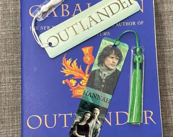May include: A blue hardcover book with the title "Outlander" by Diana Gabaldon. The book is accompanied by three bookmarks, one with the title "Outlander" in gold lettering, one with a photo of a man in a kilt, and one with a photo of a couple.