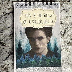 May include: Spiral-bound notepad with a printed image of a man and forest scene. The notepad has the text "THIS IS THE BILLS OF A KILLER, BELLA" in a cream-colored label. The top edge has a silver glitter strip.