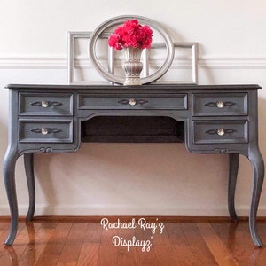 May include: A dark gray desk with six drawers and crystal knobs. The desk has curved legs and a decorative apron. A silver vase with red flowers and two silver frames are on top. The text "Rachael Ray'z Displayz" is at the bottom.