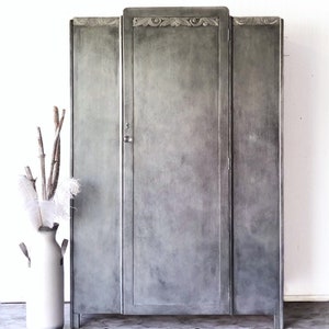 May include: A gray, three-door cabinet with decorative trim at the top. The cabinet has a keyhole and is painted in a distressed style. A white vase with branches and feathers is in front of the cabinet. The text "Rebranded by Rachael" is at the bottom.