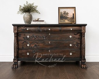 May include: A dark brown wooden dresser featuring four drawers with clear crystal knobs, intricate carved bird and floral motifs, and claw feet. The text 'rebranded by Rachael' is visible on the front. The dresser has ornate side columns.
