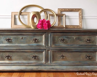 May include: A large, antique-style dresser with six drawers, painted in shades of blue and gold. The dresser features ornate hardware and is topped with gold picture frames, a vase, and pink flowers. The wood floor adds warmth.