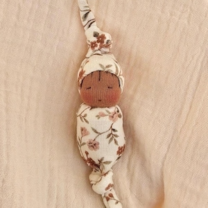 May include: A small fabric doll with a dark brown complexion, closed eyes, and rosy cheeks. The doll is wrapped in a cream-colored swaddle featuring a floral pattern of pink and brown flowers and green leaves, with a matching knotted hat and base.