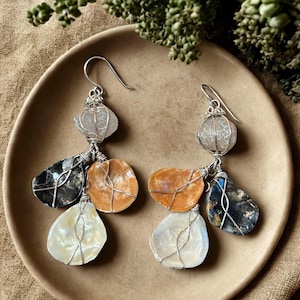 May include: A pair of dangle earrings featuring silver-toned wire-wrapped components. Each earring includes a translucent white stone at the top, with three dangling elements below in shades of iridescent white, dark gray, and warm orange.