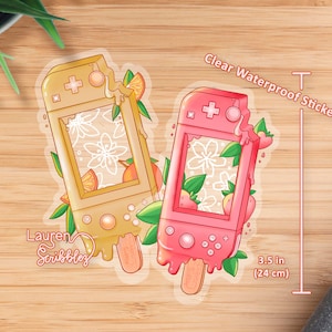 May include: Two clear waterproof stickers featuring a handheld video game console shaped like a popsicle. The left sticker is yellow with orange accents and the right sticker is pink with red accents. Both stickers have a white floral design and the text "Lauren Scribbles" is on the left sticker. The stickers are 3.5 inches (24 cm) tall.