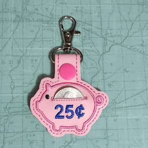 May include: Pink piggy coin purse keychain with a silver metal clasp. The purse has a slot for a quarter and the text "25¢" embroidered in blue.