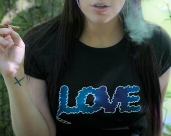 May include: A black short-sleeved t-shirt featuring the word LOVE printed in a blue and purple cloud-like graphic design. The letter E in the word LOVE is connected to a small graphic of a lit cigar at the bottom of the text design.