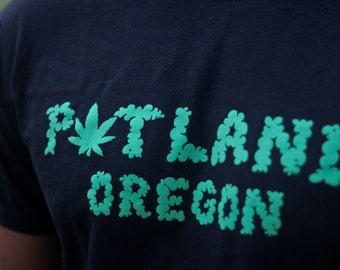 May include: A black t-shirt featuring the text "POTLAND OREGON" printed in bright green. The letter "O" in "POTLAND" is replaced by a green cannabis leaf graphic, and the letters are formed by a cloud-like, textured green pattern.