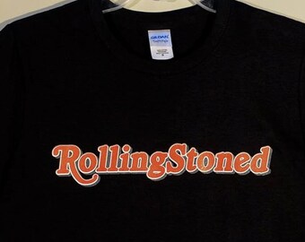 May include: A black short-sleeve t-shirt featuring the text RollingStoned printed across the chest in an orange font with a white outline. The shirt has a crew neckline and is displayed on a clear plastic hanger against a plain background.