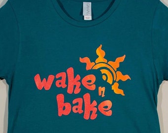 May include: A teal short-sleeved crew neck t-shirt featuring the text "wake n bake" in a bold, red-orange font. Above the text is a stylized sun graphic in a matching orange hue. The shirt has a standard fit and a simple, clean design.