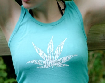May include: A turquoise sleeveless vest top featuring a white graphic print of a leaf with a zebra stripe pattern on the front, paired with white trousers. The vest top has a round neckline and a fitted silhouette, showcasing a simple design.