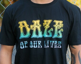 May include: A black short-sleeved t-shirt featuring the text DAZE OF OUR LIVES printed on the chest. The word DAZE is in a large, stylized font with a yellow-to-teal gradient, while the words OF OUR LIVES appear below in a smaller white font.