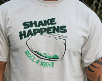 May include: A cream-colored short-sleeve t-shirt featuring a graphic print in dark green and black. The design includes the text SHAKE HAPPENS above a drawing of a small bag containing green material, with the text ROLL A JOINT below it.